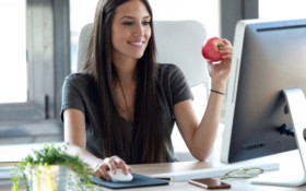 Smiling young business woman eating a red apple while working with computer in the office.