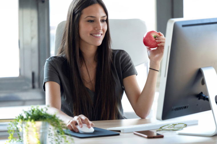 Smiling young business woman eating a red apple while working with computer in the office.
